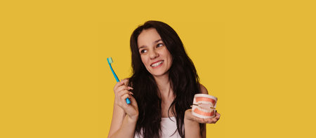 A young Caucasian brunette woman with a toothbrush and a jaw model on a yellow background. A panoramic banner with a place for your text.の写真素材