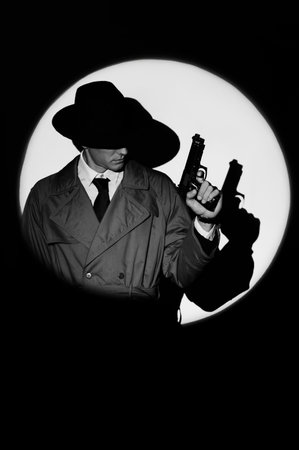 A dark silhouette of a male detective in a coat and hat with a gun in his hands in the noir style. A dramatic portrait in the style of detective films of the 1950s. The silhouette of a spy in a circle of light.の写真素材