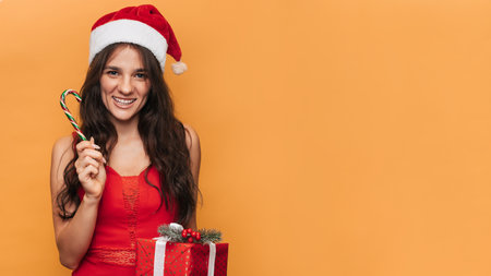 A smiling Caucasian woman in a red Santa hat with a gift box in her hands on a yellow isolated background. The concept of Christmas and New Year. A place for your text.の写真素材