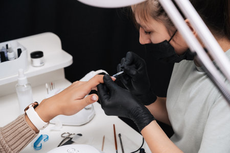 Manicure service. Close-up. Applying nail polish, leveling the surface. The manicurist paints her nails with a transparent gel polish. Well-groomed nails. Professional salon hand care.の写真素材