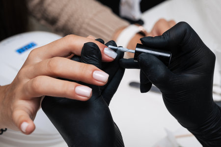 Manicure service. Close-up. Application of light varnish . The manicurist paints her nails with a transparent gel polish. Well-groomed nails. Professional salon hand care.の写真素材