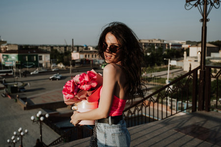 a beautiful young woman in a summer outfit, smiling happily, walking with flowers along a city streetの写真素材