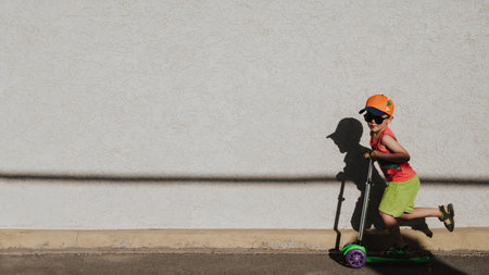 A little boy rides a scooter highlighted on a light wall. Open air. Minimalism. Panoramic banner.の写真素材