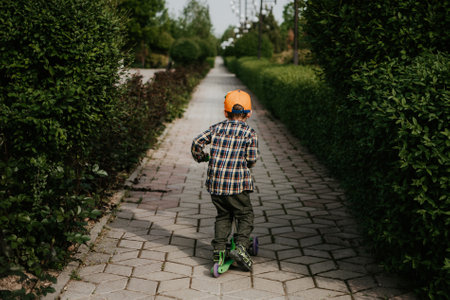 A little boy rides a scooter outdoors in a park among a green hedge. The view from the back. Minimalism.の写真素材