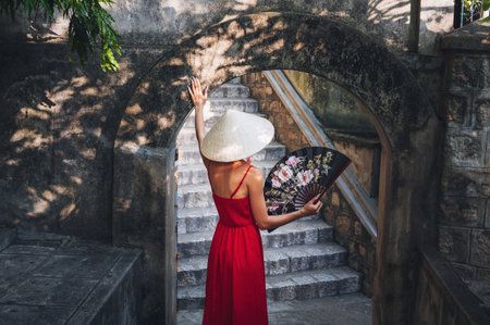 A tourist in a red dress and a traditional Vietnamese hat holding a fan in the garden of a pagoda in Asiaの写真素材