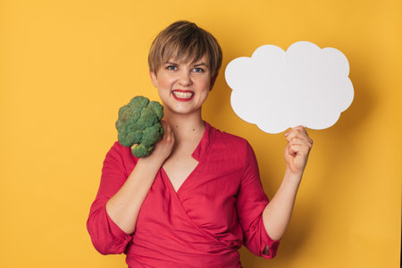 A studio shot of a young smiling woman holding fresh broccoli and a blank letterhead. The concept of healthy eating.の写真素材