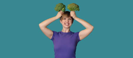 In the studio picture, a young Caucasian woman is holding fresh broccoli in her hands, and on top of her head. The concept of healthy eating. A place for your text. Panoramic banner.の写真素材