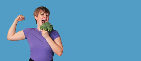 Smiling Caucasian girl with fresh broccoli in one hand and showing biceps with the other. Panoramic banner, free space.の写真素材