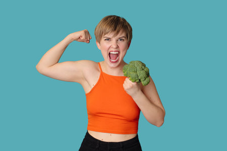 An expressive portrait of a young woman with broccoli as dumbbells on a blue background. Healthy diet, healthy lifestyle.の写真素材