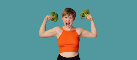 An expressive portrait of a young woman with broccoli as dumbbells on a blue background. Healthy diet, healthy lifestyle. A panoramic banner with a copy of the space.の写真素材