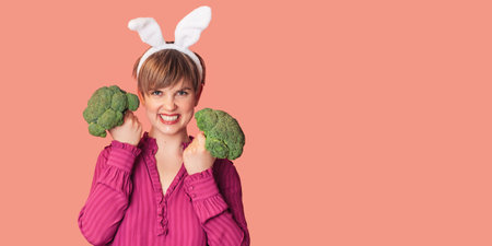 Smiling slender Caucasian girl in bunny ears with green broccoli in her hands, free space. Organic vegetablesの写真素材