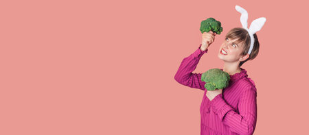Studio shot of a young Caucasian woman in bunny ears holding organic and fresh broccoli in her hands. healthy eating. A panoramic banner with copy space.の写真素材