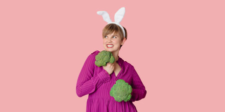 Studio shot of a young Caucasian woman in bunny ears holding organic and fresh broccoli in her hands. healthy eating. A panoramic banner with copy space.の写真素材