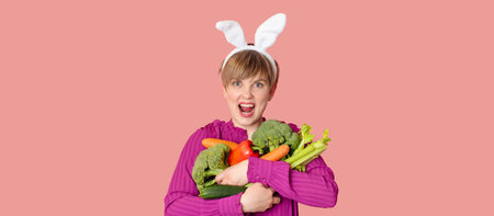 Studio shot, a young Caucasian woman in bunny ears holding a lot of organic and fresh vegetables in her hands. healthy eating.A panoramic banner with space for text.の写真素材