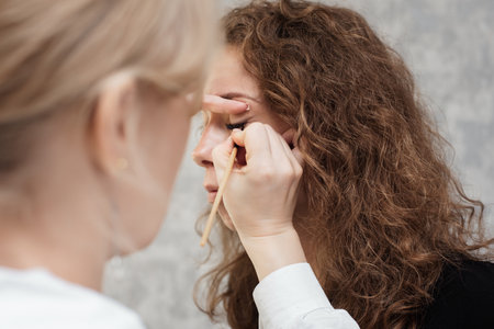 Shooting in a beauty salon. A young beautiful girl model makeup artist puts shadows on the eyelid draws a colored arrow on the eyelid.の写真素材