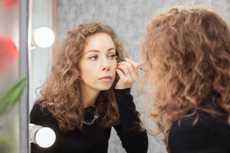 Beautiful girl is doing a makeup, retouching eyebrows while looking at the mirrorの写真素材