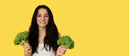 Broccoli in the hand of a slender smiling Caucasian young woman on a yellow background. healthy lifestyle. Panorama, a place for textの写真素材