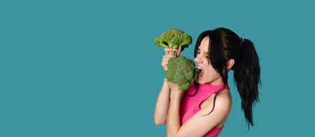 A slender smiling Caucasian young woman bites fresh broccoli on a blue background. Panorama, a place for textの写真素材