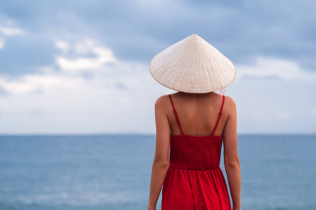 Vietnamese woman in traditional conical Non La hat and red dress on the background of the sea. View from the back.の写真素材