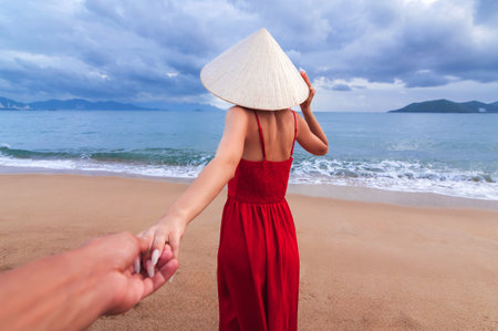 Follow me A Vietnamese woman in a traditional conical Non La hat and a red dress holds a mans hand against the background of the seaの写真素材