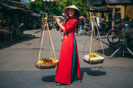 A young woman in a Vietnamese outfit carries a fruit basket near a market in the ancient city of Hoi An in Vietnam.の写真素材