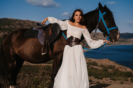 A young girl rider poses next to her horse against the backdrop of a mountain and sea landscape. The concept of riding. Artistic photography. Ready-made cover for books and magazines.の写真素材