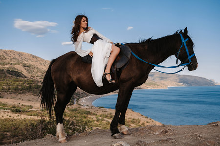 A young lady rider in a white dress poses in the saddle on a horse leaning back against a beautiful mountain landscape. The concept of romance, fairy tales. Example of a finished book coverの写真素材