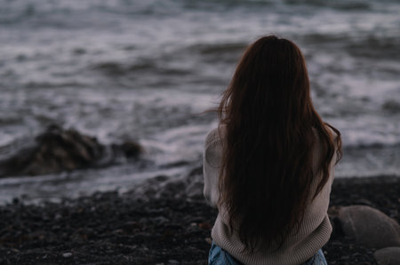 A young Caucasian girl is sitting on a rock near the seashore, and looks at the horizon. Gray, cool, inclement weather.の写真素材