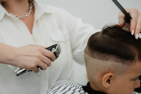 A female hairdresser cuts the back of a little boys head with a clipper. A professional hairdresser and a child client in a modern barbershop.の写真素材