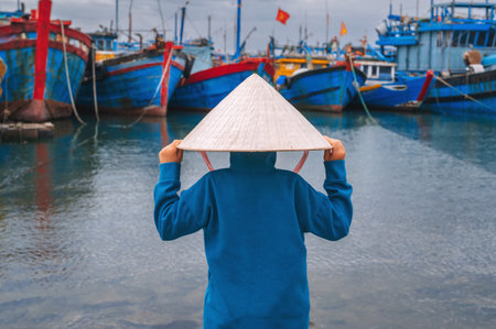 A child traveler in a traditional Vietnamese hat, inspecting traditional wooden boats in the port of Da Nang.の写真素材