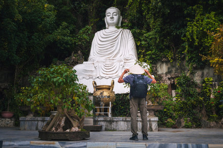 A male traveler in a Vietnamese hat at the white marble Buddha statue on the Marble Mountains in Da Nang.の写真素材