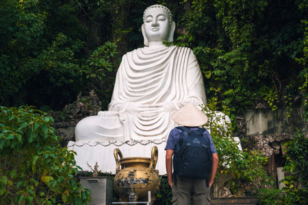 A male traveler in a Vietnamese hat at the white marble Buddha statue on the Marble Mountains in Da Nang.の写真素材