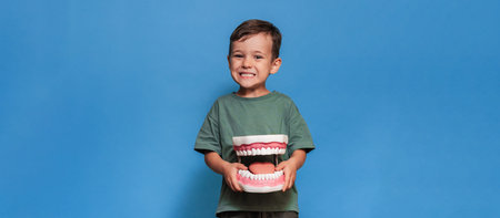 A smiling boy with healthy teeth holds a large jaw in his hands on a blue isolated background. Oral...