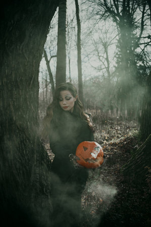 A young woman in a Gothic gloomy image of a witch surrounded by smoke, a pumpkin in her hands. Halloween costume. Vertical photo.の写真素材