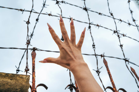 womens hand behind a barbed wire fence against the sky.