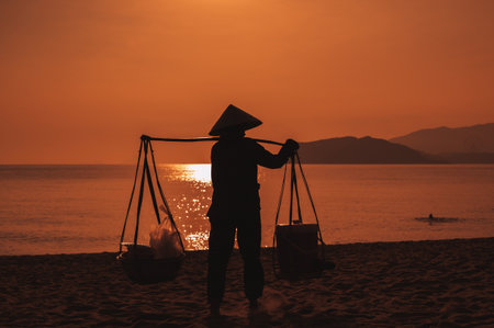 Silhouette of a woman in a traditional Vietnamese hat carrying goods along the seashore at dawn.の写真素材