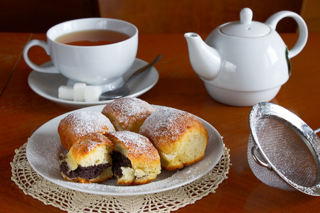 Sugared poppy seed buns on a plate with a cup of tea and porcelain pot on a wooden table. A sweet breakfast with lace placemats.の写真素材