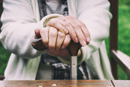 Senior woman sitting outside on a wooden bench and rests. Hands of a woman pensioner holding a walking stick.の写真素材