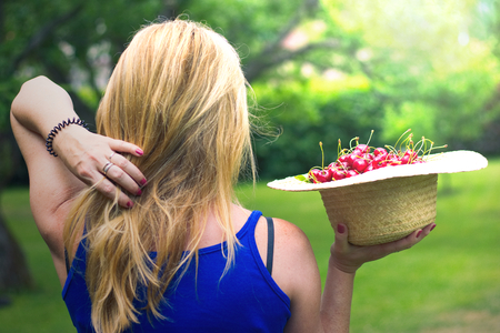Young woman holding a straw hat full of ripe sweet cherries. Summer harvest of red cherries in the garden.の写真素材