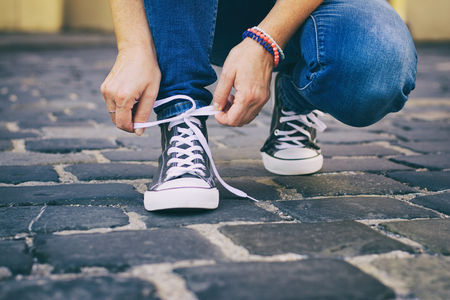 A woman binds laces on her sneakers in old town. Woman wearing jeans and trendy footwear. の写真素材