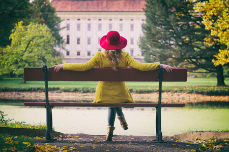 Fashion woman with red hat sitting on a bench in park and enjoying view on a castle, autumn season.の写真素材