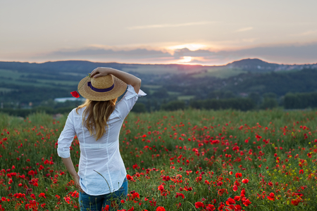 Young woman standing in poppy field dressed in jeans and white shirt watching sunset. Romantic portrait of a woman in straw hat on a meadow.の写真素材