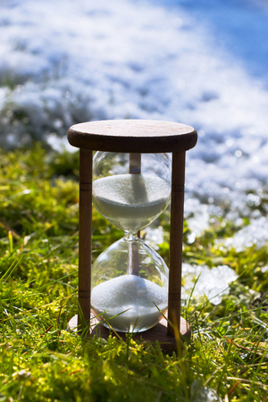 Hourglass as a symbol of climate change. Global warming. Spring is coming. Ending winter. Hour-glass in the grass and melting snow.の写真素材