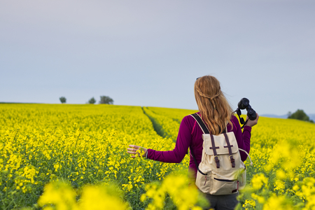 Hiking woman watching landscape with binoculars. Ornithologist in the middle of blossoming rapeseed field at spring. の写真素材