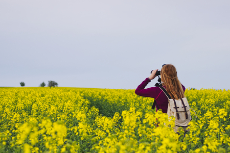 Hiking woman watching landscape with binoculars. Ornithologist in the middle of blossoming rapeseed field at spring. Bird watching. の写真素材