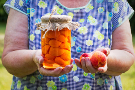 Senior woman holding glass jar of preserved apricots and ripe apricot in her hands. Old female cook in garden.の写真素材