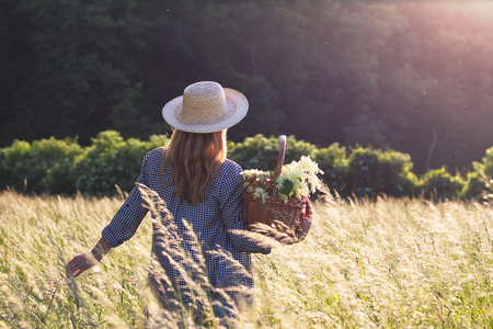 Woman with straw hat is holding wicker basket and standing in meadow. Elderberry harvest. Tranquility scenery with woman in sunlight.の写真素材