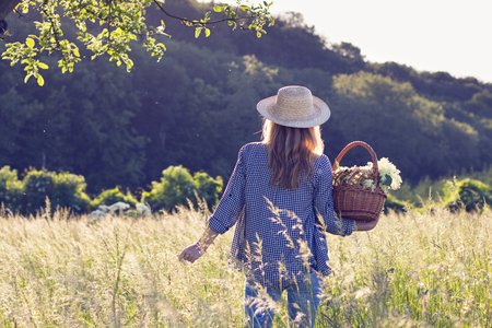 Woman with straw hat is holding wicker basket and standing in meadow. Elderberry harvest. Tranquility scenery with woman in sunlight.の写真素材