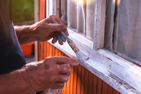 Senior man painting wooden windows using paintbrush. Repairing exterior of old house. Close-up view on old hand holding paintbrush.の写真素材