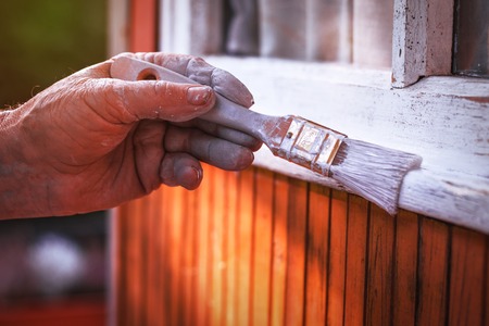 Senior man painting wooden windows using paintbrush. Repairing exterior of old house. Close-up view on old hand holding paintbrush.の写真素材